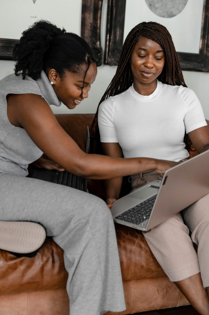 Two women smiling and collaborating on a laptop while sitting comfortably at home.